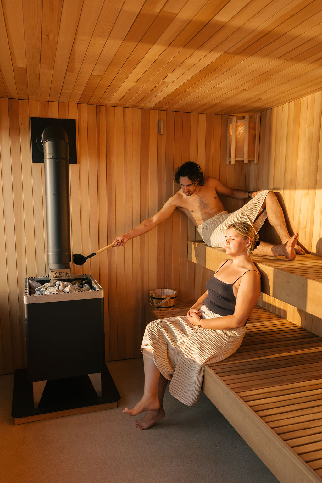 Two guests in the cedar sauna — one ladles water onto the stove while the other leans back on the lower bench