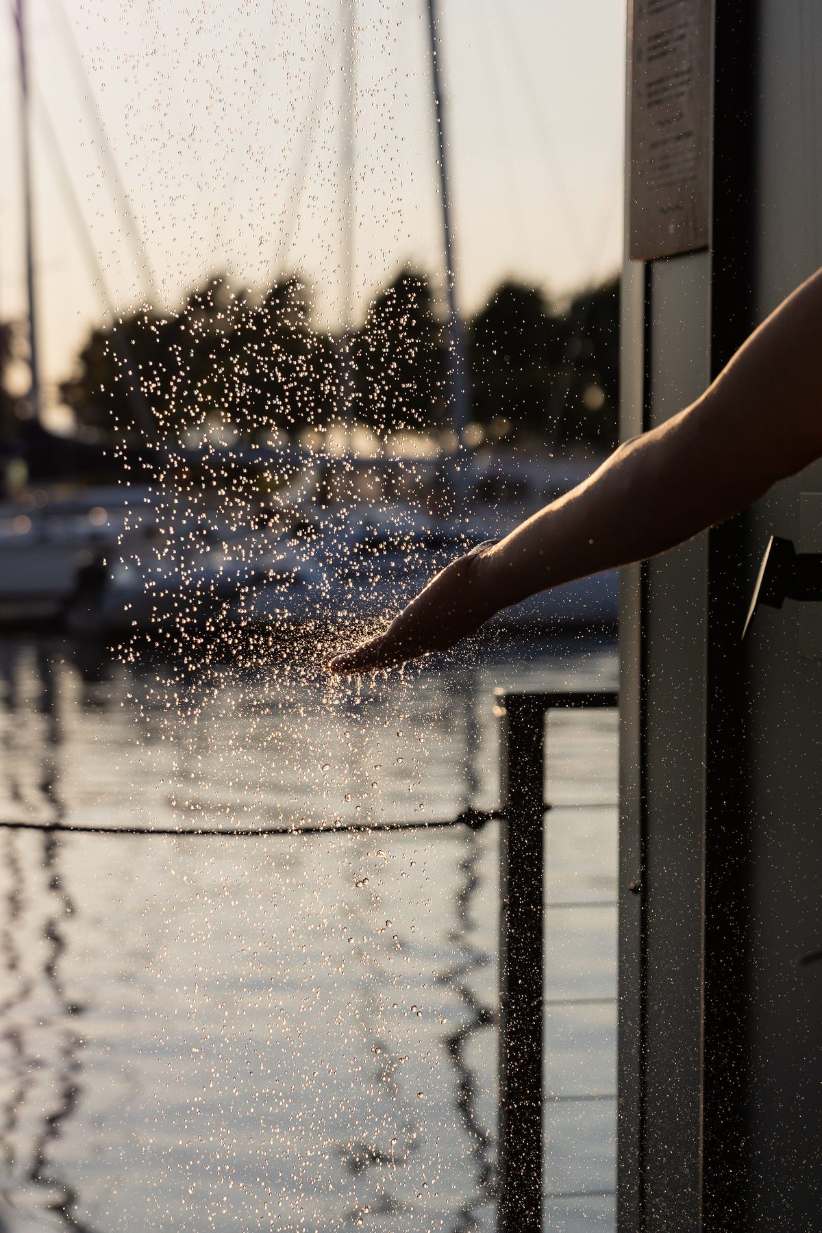 A hand reaching into the outdoor shower spray, marina sailboats glowing behind