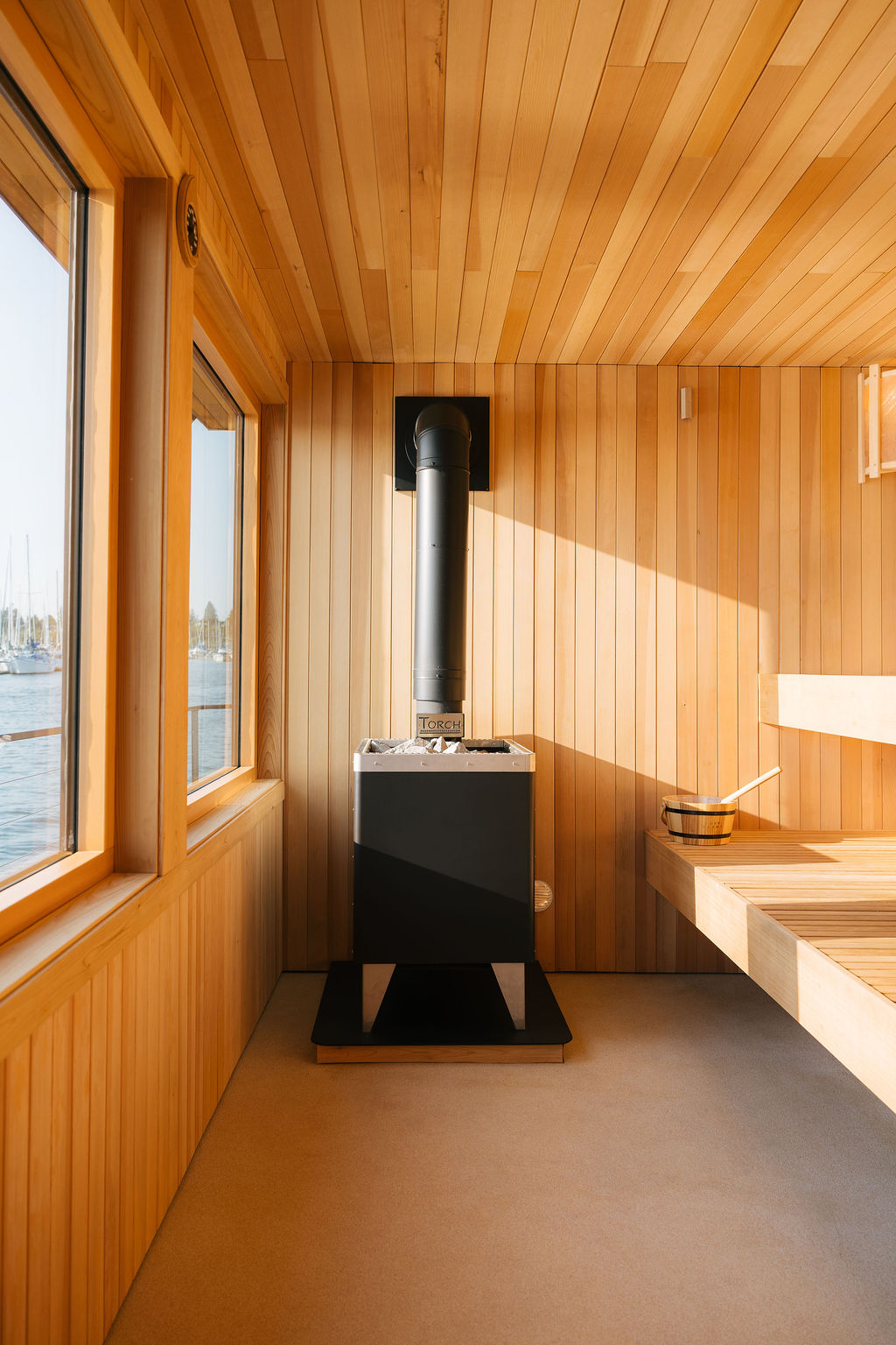 The full cedar sauna interior — stove at the far end, benches along one side, windows onto the bay