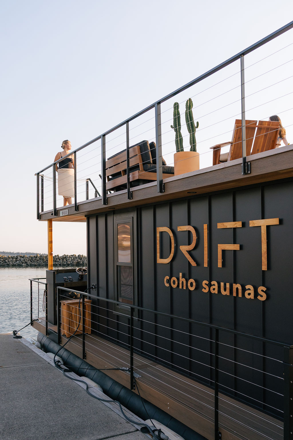 The black Drift sauna boat at the dock, guests on the rooftop deck above the cedar Drift lettering